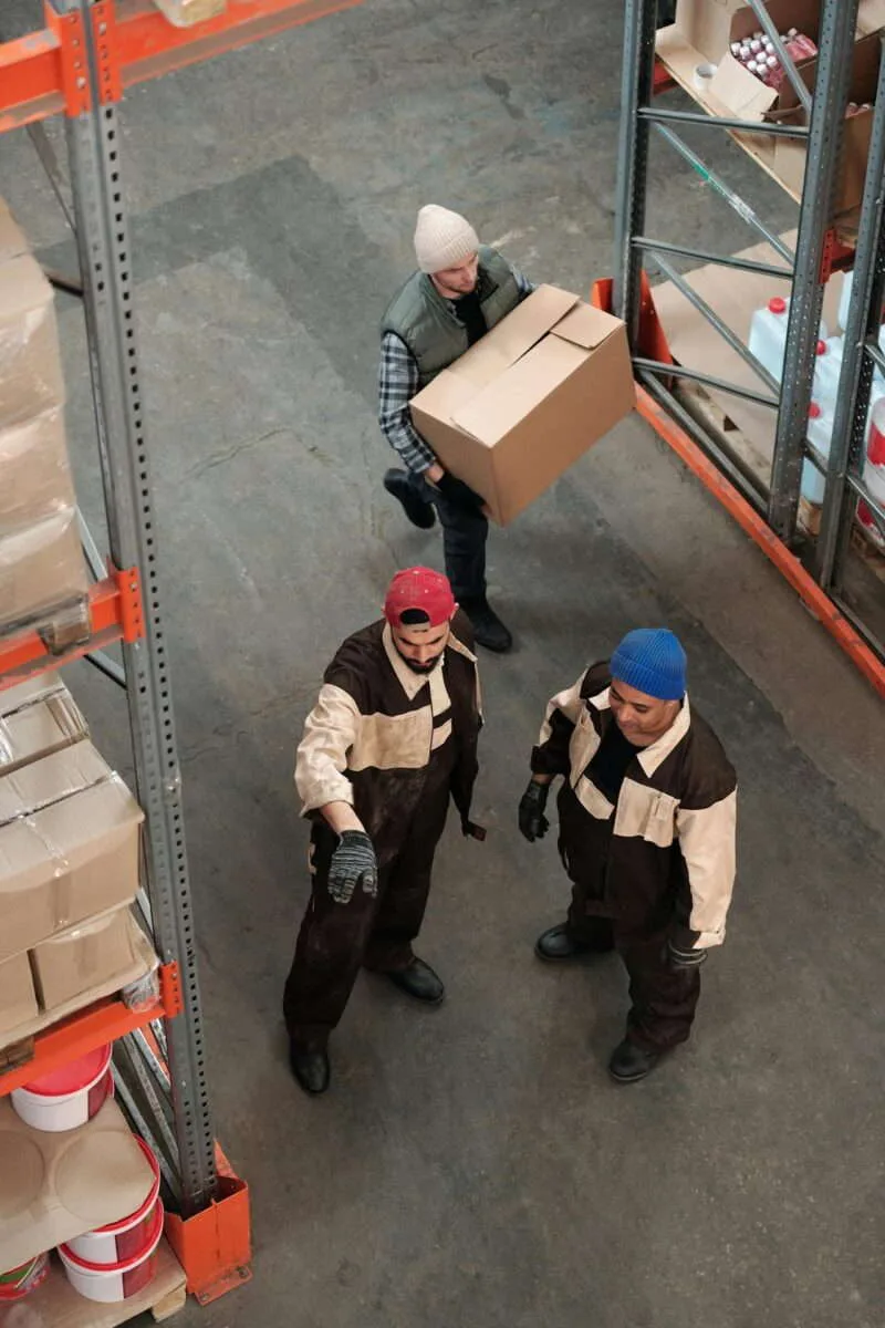 Overhead view of three warehouse workers in an aisle between orange pallet racks, one carrying a cardboard box