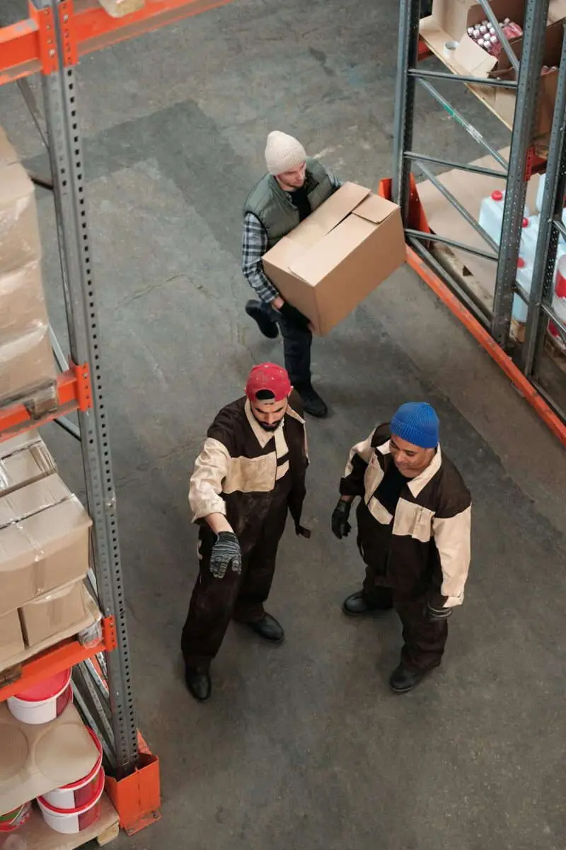 Overhead view of three warehouse workers between orange pallet racks, one carrying a cardboard box while two others review the next shipment.